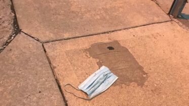 Shows a light blue paper surgical mask that was used against the coronavirus. It's been discarded on the sidewalk